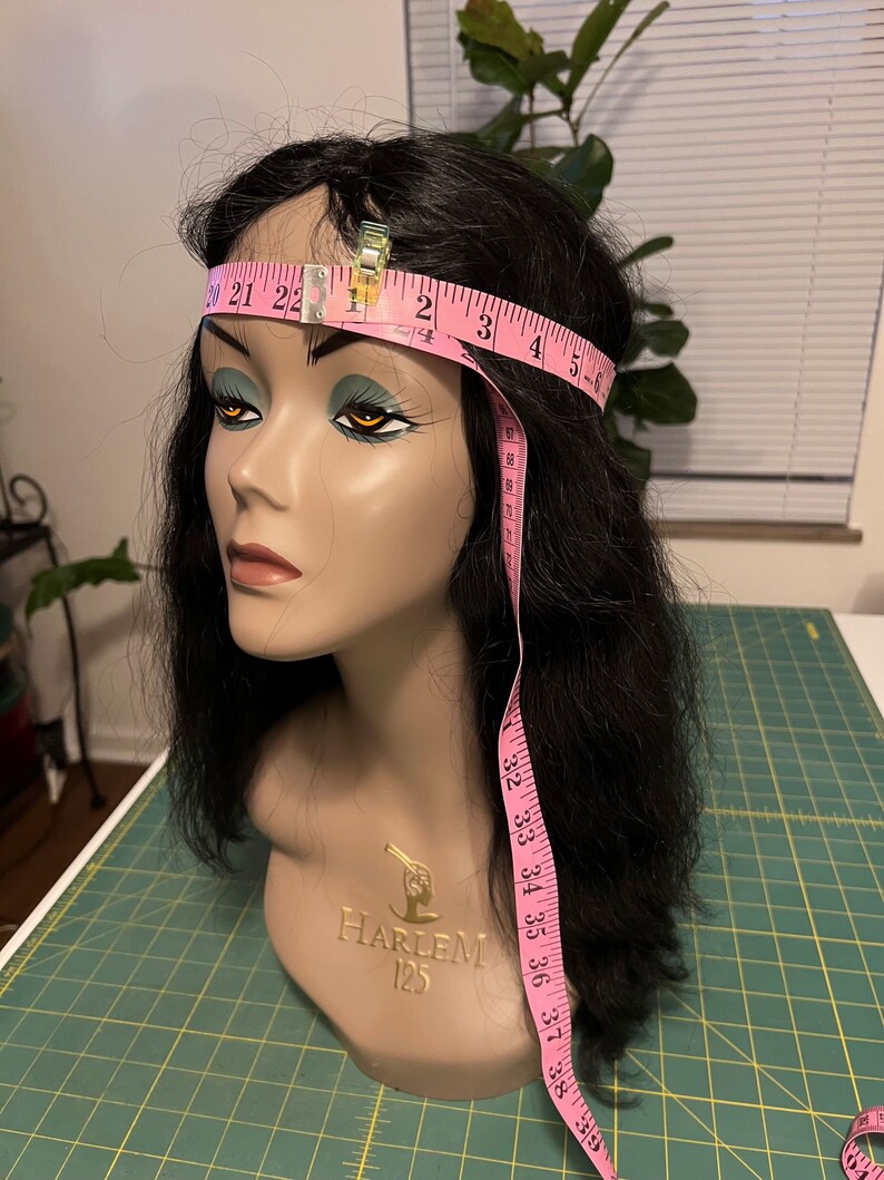 May include: A mannequin head with black hair and a pink measuring tape wrapped around the head. The tape is held in place with a metal clip. The mannequin has the words "HARLEM 125" on the neck.