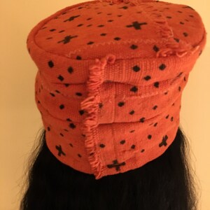 May include: An orange fabric hat with black cross and dot patterns. The hat has a rounded top and a layered, banded design. The edges of the hat have a fringed trim. The hat is worn over black hair.