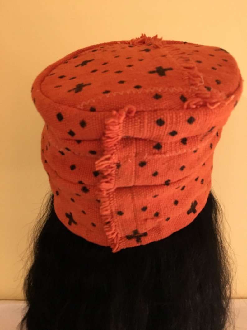 May include: An orange fabric hat with black cross and dot patterns. The hat has a rounded top and a layered, banded design. The edges of the hat have a fringed trim. The hat is worn over black hair.
