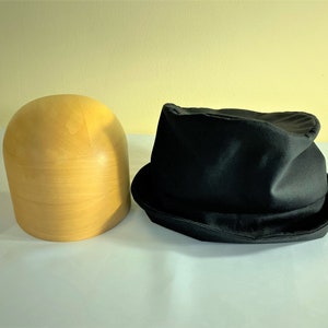 May include: A black fedora hat with a slightly crumpled crown sits next to a light-colored wooden hat block. The hat has a classic brim and a dark, matte finish. The hat block is smooth and cylindrical.