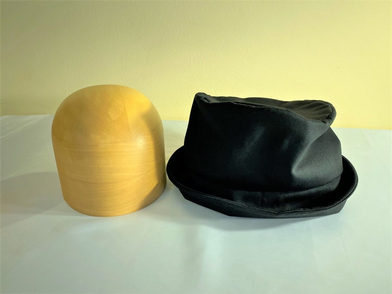 May include: A black fedora hat with a slightly crumpled crown sits next to a light-colored wooden hat block. The hat has a classic brim and a dark, matte finish. The hat block is smooth and cylindrical.