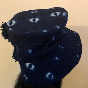 Puede incluir: Un gorro de punto azul marino con un patr&oacute;n &uacute;nico de formas blancas en forma de ojo. El gorro tiene un borde con flecos y un borde doblado. El gorro est&aacute; hecho de una tela suave y texturizada, y es un accesorio elegante para el clima fr&iacute;o.