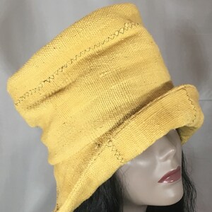 May include: A yellow, textured hat with a brim and a tall crown. The hat has a stitched design along the seams. The hat is displayed on a mannequin head with dark hair.