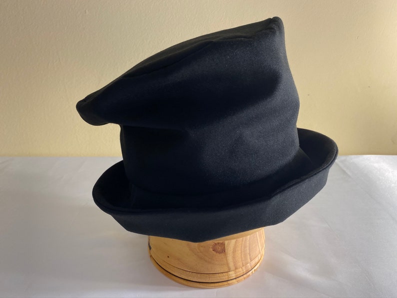May include: A black hat with a unique, asymmetrical design. The hat has a wide brim and a tall, pointed crown. It is displayed on a wooden hat stand. The hat appears to be made of a soft, felt-like material.
