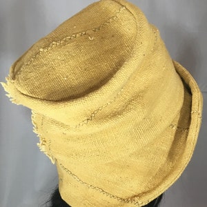 May include: Mustard yellow hat with a brim and a slightly pointed crown. The hat is made of a woven fabric with visible texture and is stitched with dark thread. The brim has a frayed edge.