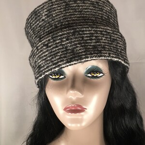 May include: A black and white patterned cloche hat. The hat has a wide brim and a rounded crown. The hat is made of a textured fabric and has a white trim along the bottom edge. The hat is displayed on a mannequin.