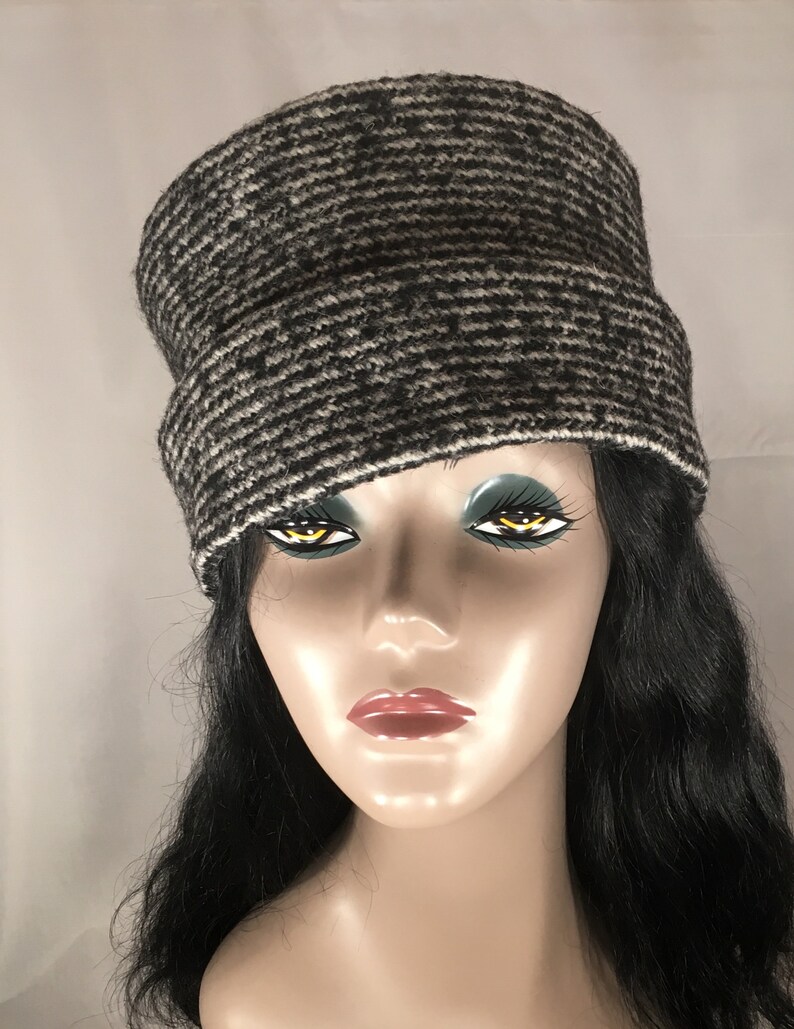 May include: A black and white patterned cloche hat. The hat has a wide brim and a rounded crown. The hat is made of a textured fabric and has a white trim along the bottom edge. The hat is displayed on a mannequin.