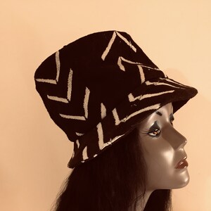 May include: A black bucket hat featuring a white zigzag design. The hat is crafted from a textured material and has a gently sloping brim. The hat is shown on a mannequin head.