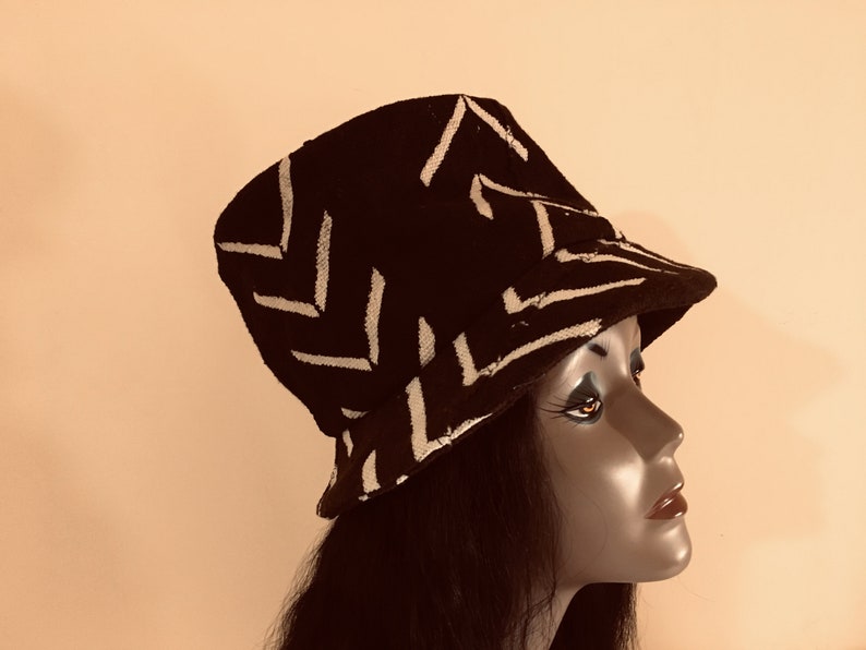 May include: A black bucket hat featuring a white zigzag design. The hat is crafted from a textured material and has a gently sloping brim. The hat is shown on a mannequin head.