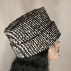 May include: A black and white patterned hat with a brim. The hat is made of a textured fabric and has a cylindrical shape. The hat is displayed on a mannequin head with long black hair.