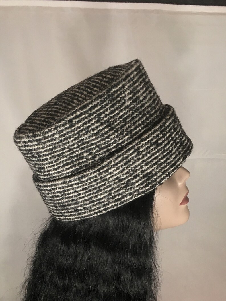 May include: A black and white patterned hat with a brim. The hat is made of a textured fabric and has a cylindrical shape. The hat is displayed on a mannequin head with long black hair.