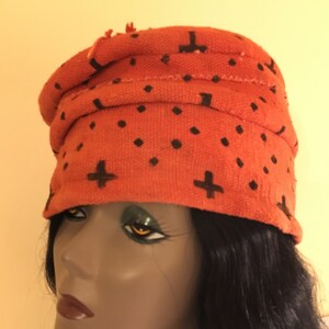 May include: An orange fabric head wrap with black cross and dot patterns. The head wrap is wrapped around a mannequin head. The head wrap is a turban style with a rolled brim.