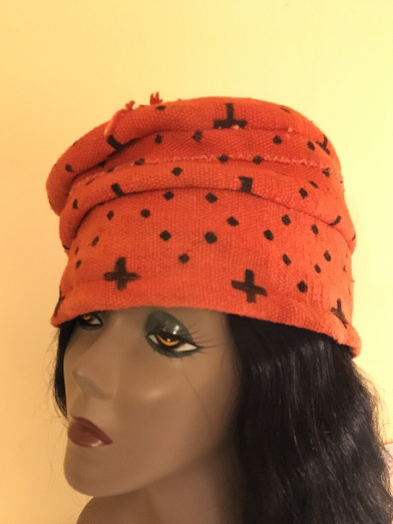 May include: An orange fabric head wrap with black cross and dot patterns. The head wrap is wrapped around a mannequin head. The head wrap is a turban style with a rolled brim.
