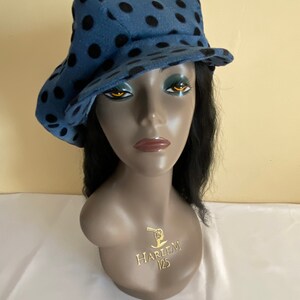 May include: A blue hat with black polka dots. The hat is displayed on a mannequin head. The hat has a brim that is slightly turned up on one side. The mannequin has black hair and a gold emblem that says "HARLEM 125".