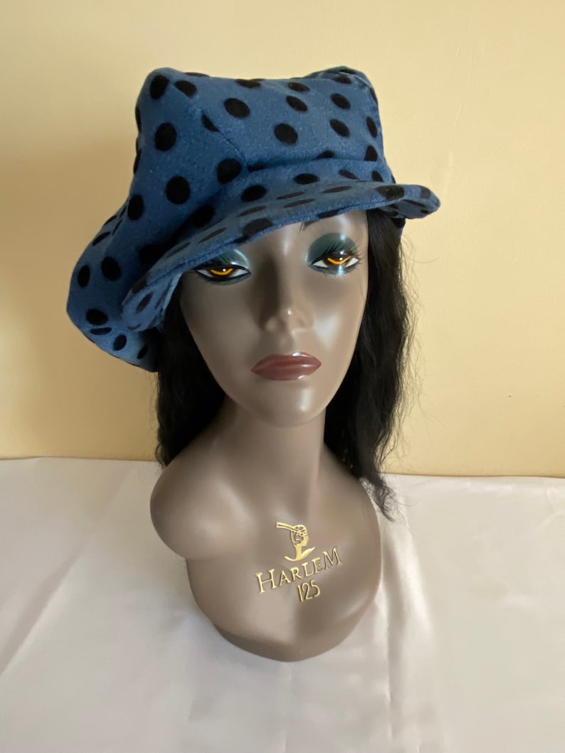 May include: A blue hat with black polka dots. The hat is displayed on a mannequin head. The hat has a brim that is slightly turned up on one side. The mannequin has black hair and a gold emblem that says "HARLEM 125".
