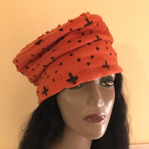 May include: An orange turban-style hat with a repeating pattern of black cross and dot shapes. The hat is made of a textured fabric and has a rolled design. The hat is worn by a mannequin with long black hair.