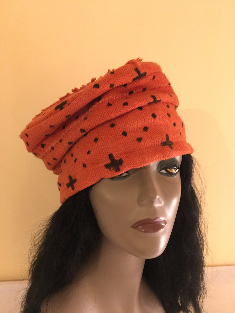 May include: An orange turban-style hat with a repeating pattern of black cross and dot shapes. The hat is made of a textured fabric and has a rolled design. The hat is worn by a mannequin with long black hair.