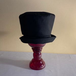 May include: A black top hat with a cylindrical crown and a flat brim. The hat is displayed on a red glass pedestal. The hat's classic design is suitable for formal events or costume parties.