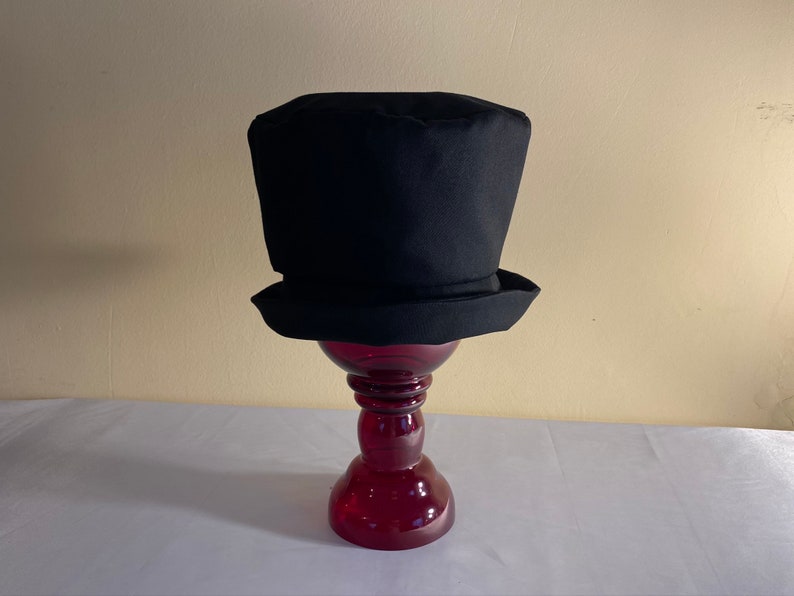 May include: A black top hat with a cylindrical crown and a flat brim. The hat is displayed on a red glass pedestal. The hat's classic design is suitable for formal events or costume parties.