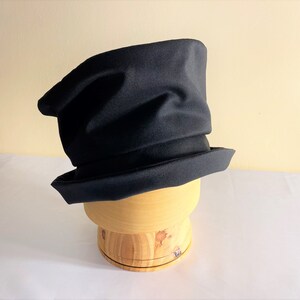 May include: A black hat with a unique, asymmetrical design. The hat has a wide brim and a tall, slightly crumpled crown. It is displayed on a wooden hat form, showcasing its shape and structure. The hat's dark color and distinctive shape make it a stylish accessory.