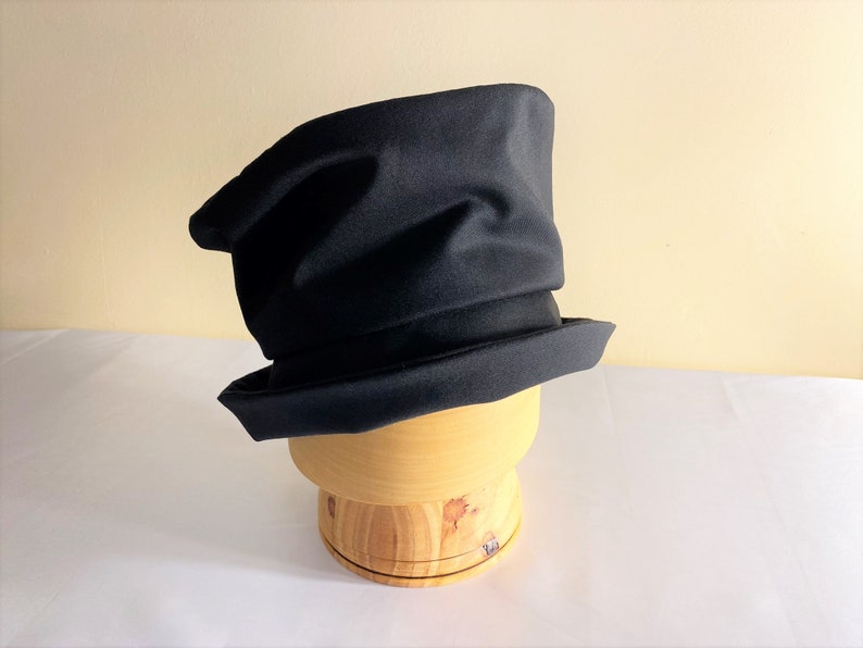 May include: A black hat with a unique, asymmetrical design. The hat has a wide brim and a tall, slightly crumpled crown. It is displayed on a wooden hat form, showcasing its shape and structure. The hat's dark color and distinctive shape make it a stylish accessory.