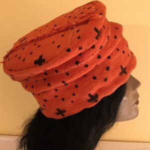 May include: An orange fabric hat with a layered design and black cross and dot patterns. The hat has a frayed edge and is worn on a mannequin head with long black hair. The hat is a unique fashion accessory.