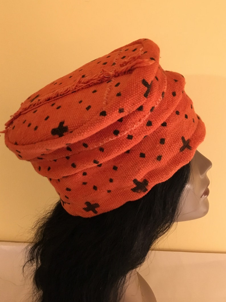 May include: An orange fabric hat with a layered design and black cross and dot patterns. The hat has a frayed edge and is worn on a mannequin head with long black hair. The hat is a unique fashion accessory.