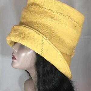 May include: A yellow hat with a wide brim and a tall crown. The hat is made of a woven fabric and features visible stitching along the seams. The hat is shown on a mannequin head with black hair.
