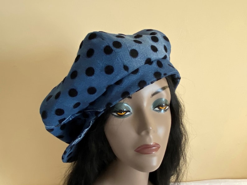 May include: A blue denim beret-style hat with large black polka dots. The hat has a brim and a rounded crown. The hat is worn by a mannequin with black hair and a neutral background.