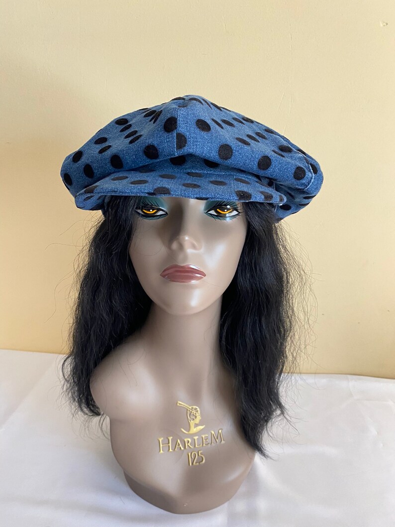 May include: A denim newsboy cap with black polka dots. The hat is displayed on a mannequin head with black hair. The mannequin has the text "HARLEM 125" on the chest.