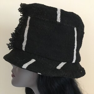 May include: Black bucket hat with white vertical and diagonal stripes. The hat has a frayed edge and a small brim. The hat is made of a woven fabric. The hat is on a mannequin head.