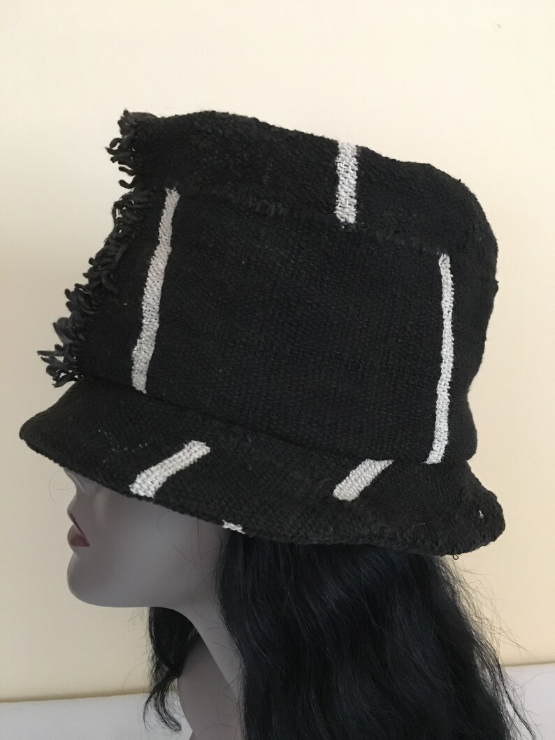 May include: Black bucket hat with white vertical and diagonal stripes. The hat has a frayed edge and a small brim. The hat is made of a woven fabric. The hat is on a mannequin head.