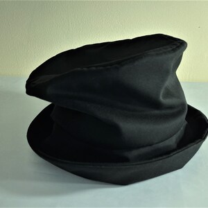 May include: A black hat with a wide brim and a tall, slightly crumpled crown. The hat is made of a soft, dark fabric and appears to be a costume or theatrical accessory. The brim is angled, and the crown has a unique, folded shape.