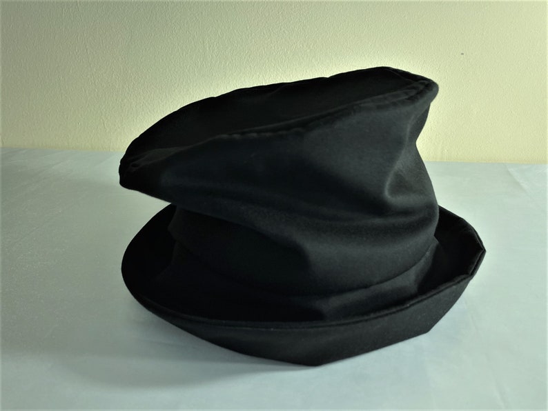 May include: A black hat with a wide brim and a tall, slightly crumpled crown. The hat is made of a soft, dark fabric and appears to be a costume or theatrical accessory. The brim is angled, and the crown has a unique, folded shape.
