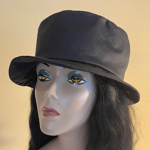 Puede incluir: Un sombrero cloche negro con una corona redondeada y un ala ligeramente curvada. El sombrero está hecho de un material liso, posiblemente satinado. El sombrero se muestra en una cabeza de maniquí con cabello oscuro.