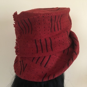 K&ouml;nnte beinhalten: Ein roter Hut mit schwarzen vertikalen Streifenmustern und Fransen. Der Hut besteht aus einem gewebten Stoff und hat eine zylindrische Form. Der Hut wird auf dem Kopf einer Person getragen.