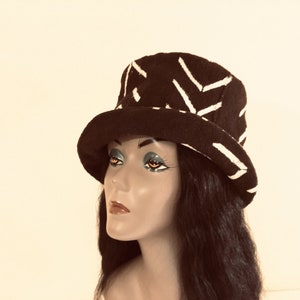 May include: A dark hat featuring a white geometric design. The hat has a rounded crown and a wide brim. It is displayed on a mannequin head with long black hair. This hat is a fashion accessory.
