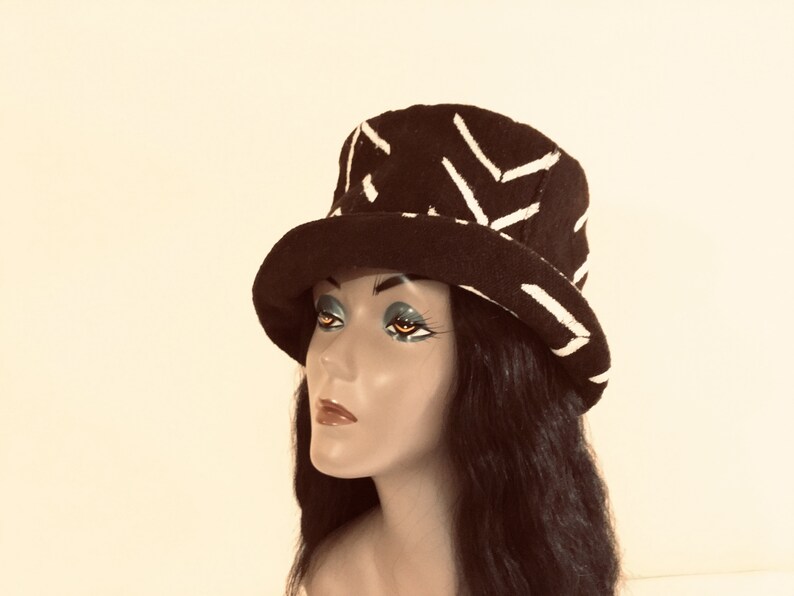 May include: A dark hat featuring a white geometric design. The hat has a rounded crown and a wide brim. It is displayed on a mannequin head with long black hair. This hat is a fashion accessory.