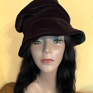 May include: A black velvet hat with a unique, asymmetrical design. The hat has a soft, draped appearance and a wide brim. The hat is displayed on a mannequin head with long, black hair. The mannequin has the text "HARLEM 125" on its chest.