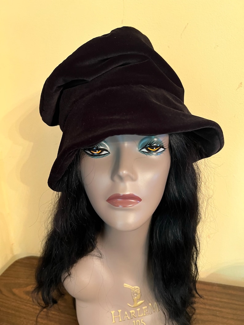 May include: A black velvet hat with a unique, asymmetrical design. The hat has a soft, draped appearance and a wide brim. The hat is displayed on a mannequin head with long, black hair. The mannequin has the text "HARLEM 125" on its chest.