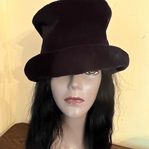 May include: A dark purple top hat with a wide brim. The hat is made of a soft material and is displayed on a mannequin head with long black hair. The mannequin has a gold necklace that says "Harlem 125".