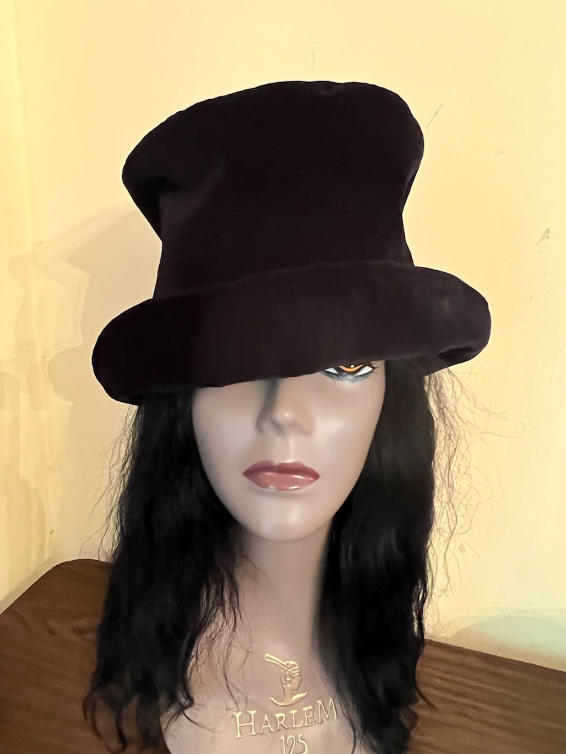 May include: A dark purple top hat with a wide brim. The hat is made of a soft material and is displayed on a mannequin head with long black hair. The mannequin has a gold necklace that says "Harlem 125".