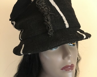 May include: A black knitted hat with white stripes and fringing. The hat features a flared brim and a rounded crown. The hat is made from a textured material and has a casual, bohemian style.