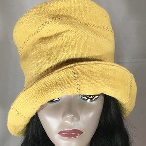 May include: A yellow bucket hat with a wide brim. The hat is made of a textured fabric and features visible stitching details. The hat is displayed on a mannequin head with black hair.