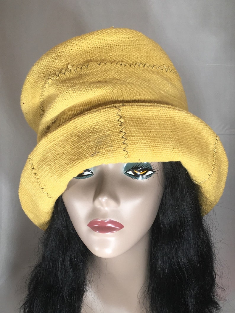May include: A yellow bucket hat with a wide brim. The hat is made of a textured fabric and features visible stitching details. The hat is displayed on a mannequin head with black hair and red lipstick.