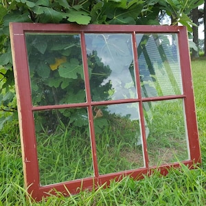 May include: A red wooden window frame with six panes of glass. The window is lying on the ground in a grassy area.