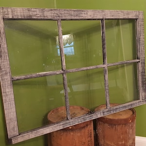 May include: A rustic, rectangular window frame with six panes of glass. The frame is made of weathered wood with a gray and white finish. The window is propped up on two brown, cylindrical barrels against a green wall.