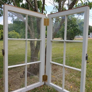 May include: Two white painted wooden window panes with brass hinges. The panes are divided into nine squares with clear glass. The windows are standing on a grassy lawn.