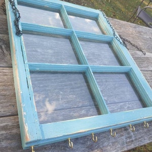 May include: A repurposed, rectangular window frame painted in a distressed light blue. The frame is divided into six panes of glass. It has a dark chain for hanging and small gold hooks along the bottom edge for displaying items.