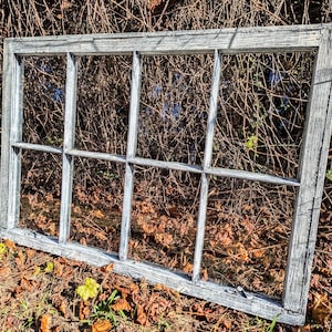 May include: A weathered white wood window frame with nine panes of glass. The window is lying on the ground with fallen leaves around it.